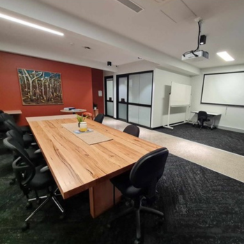 Modern conference room with a large wooden table, black chairs, whiteboard, projector, and artwork on a red accent wall.